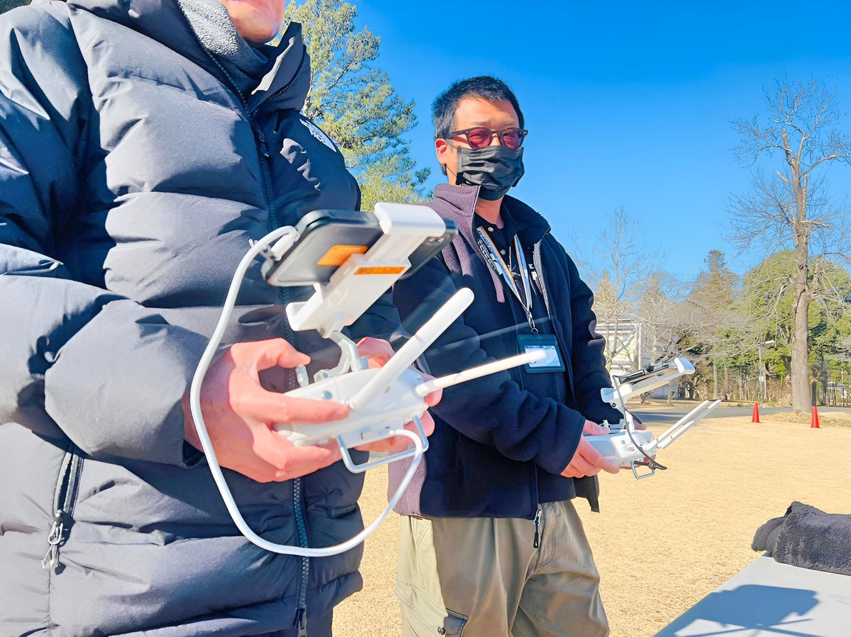 屋外訓練場で送信機を操作しながらドローンの飛行状況を確認する講師と受講者の様子