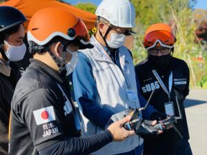 （公認）DSI ドローン・ビジネススクール茨城にて、ヘルメットを着用した受講生と講師が送信機を使い実技講習を行っている様子